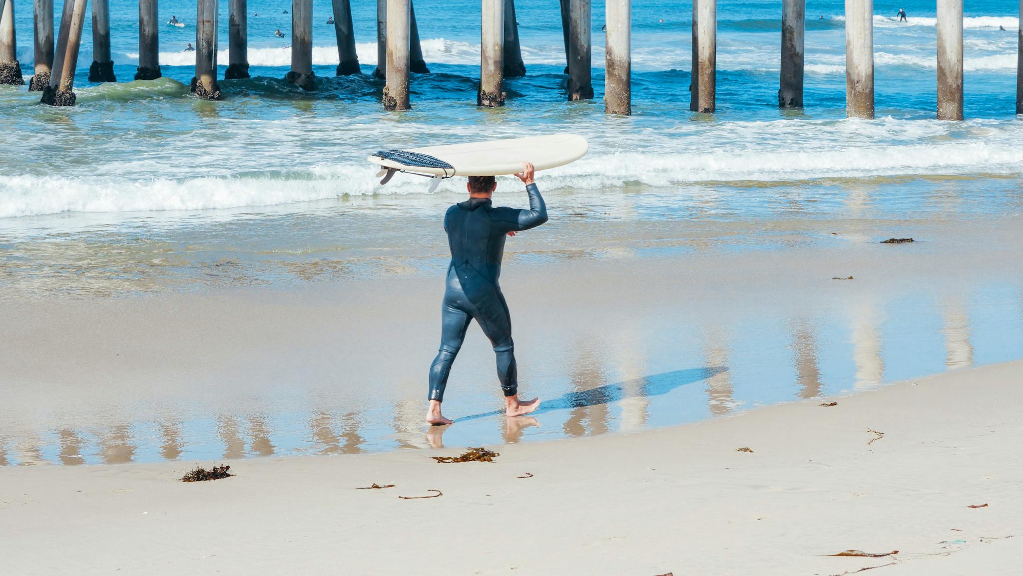 A surfer carrying a surfboard on a sandy shore by a pier in California.