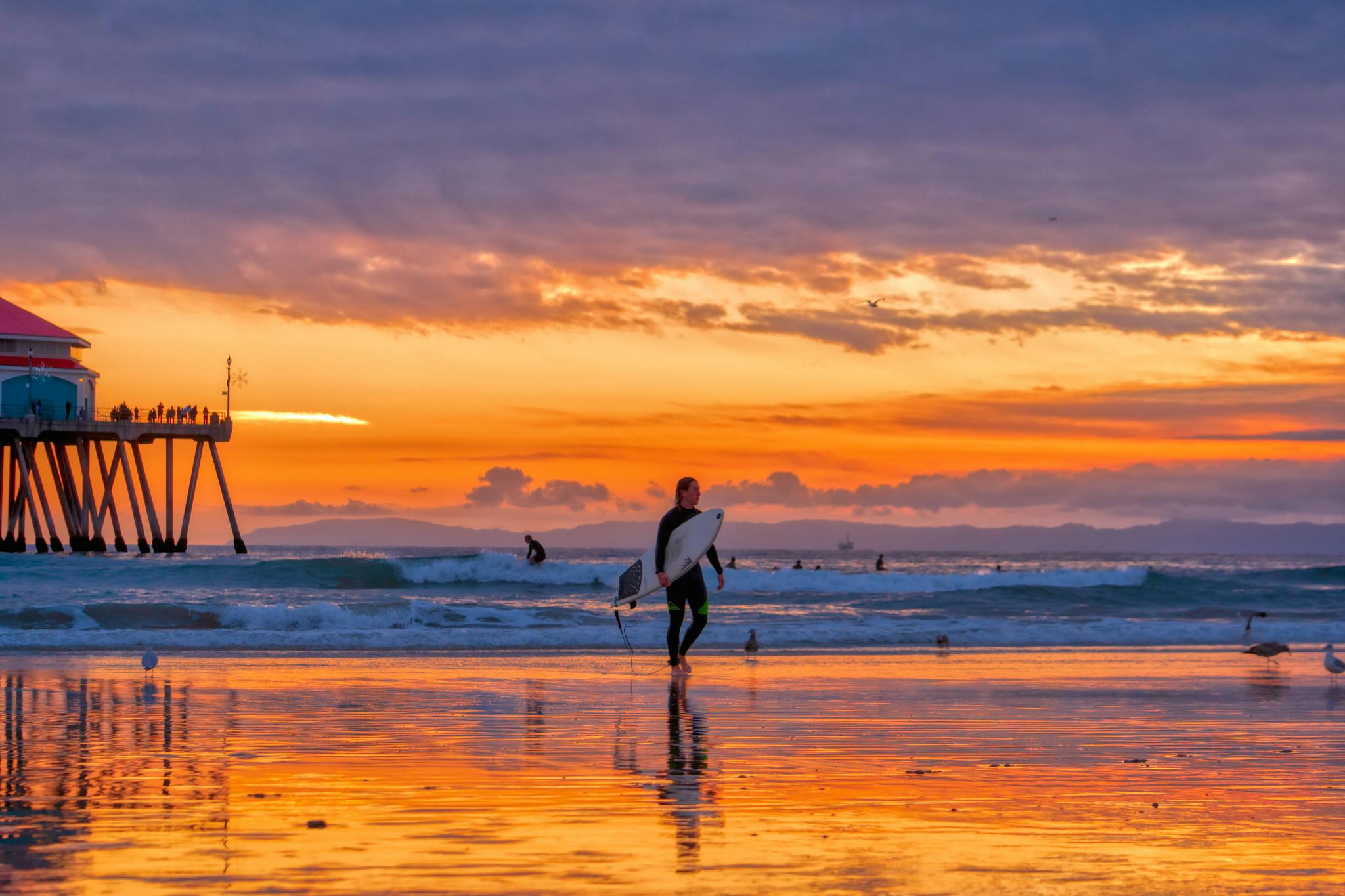 A surfer walks with their board on the reflective sands of Huntington Beach at sunset.